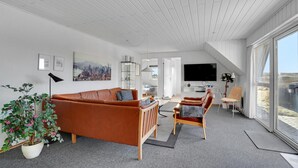 Una sala de estar moderna con techo alto, un ventanal grande, un sofá, un sillón, un televisor y una pintura de un paisaje urbano.