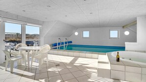 Zona de piscina interior iluminada por el sol con una mesa y sillas, un jacuzzi y vistas al paisaje a través de amplias ventanas.