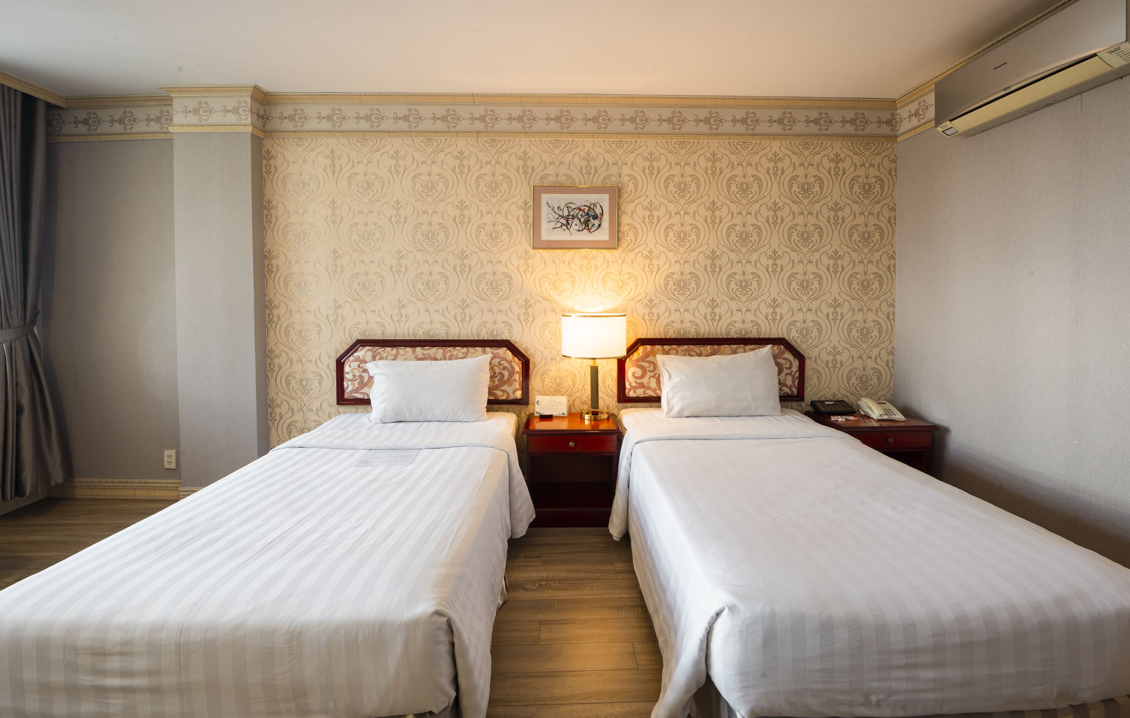 Quarto de hotel com duas camas de solteiro, uma mesa de cabeceira com abajur, um telefone e um quadro na parede.
