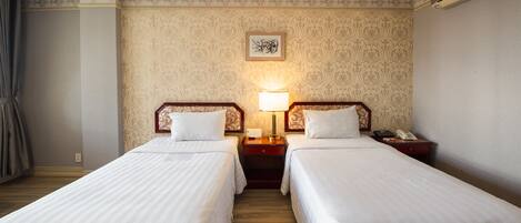 Quarto de hotel com duas camas de solteiro, uma mesa de cabeceira com abajur, um telefone e um quadro na parede.
