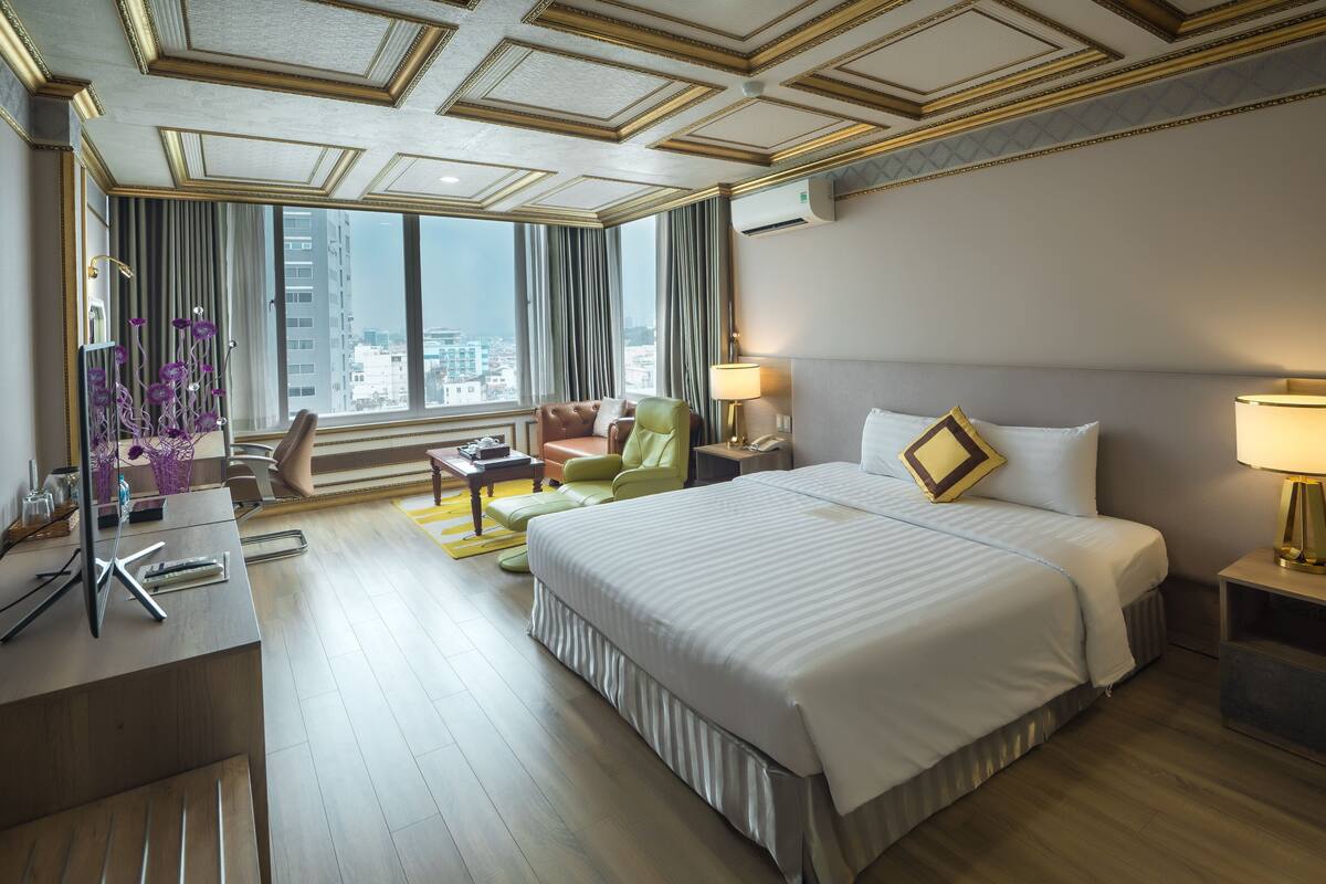 Une chambre d’hôtel avec un grand lit, un bureau équipé d’un ordinateur, une chaise, une lampe et une vue sur la ville grâce à de grandes fenêtres.