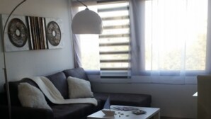 Quarto moderno com uma cama grande, um sofá, uma mesinha e uma cadeira. Há uma janela com persianas e uma ventilação no teto.