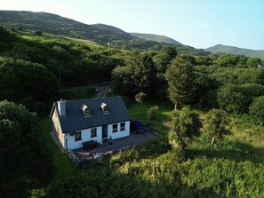 A white house with a sloped roof, a paved driveway, and a small garden area with a bench and a table. - Traditional style Cottage in a quiet location close to the Kenmare Bay (Coad Caherdaniel)