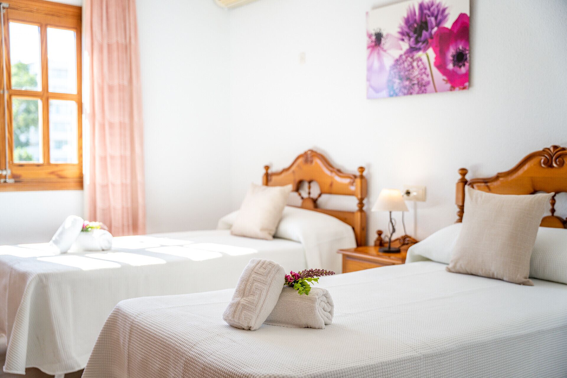 A hotel room with two beds, each with a wooden headboard, white bedding, and a small bedside table with a lamp. A window with pink curtains is visible, and a floral artwork hangs on the wall.