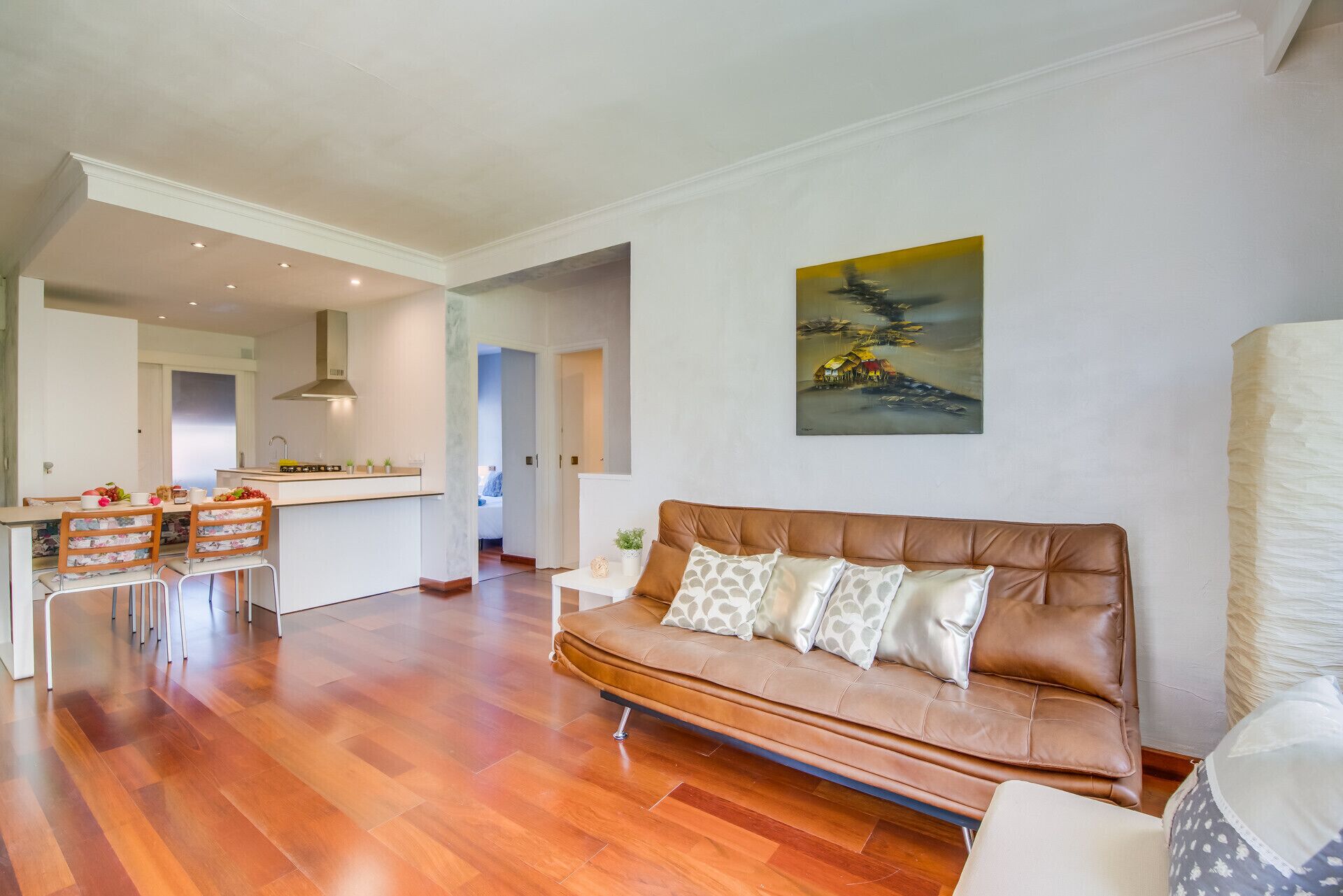 Een moderne woonkamer met een bruine leren bank, een eethoek met twee stoelen, een keuken op de achtergrond en een schilderij aan de muur.