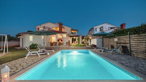 Ein Swimmingpool mit Liegestühlen und Sonnenschirmen, umgeben von einer Steinterrasse und einem Holzzaun.