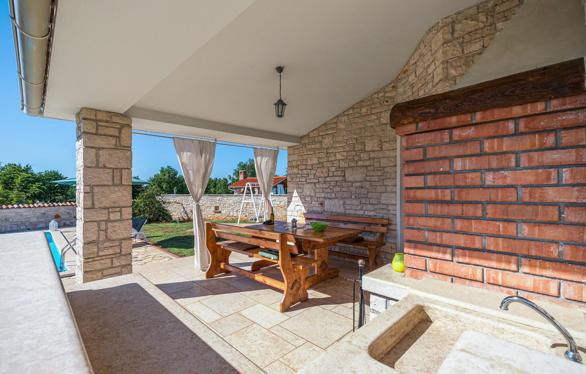 Une terrasse extérieure couverte, avec une table et des bancs en bois, des murs en pierre et une vue sur un jardin et des bâtiments en arrière-plan.