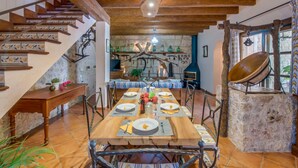 Un comedor rústico con una mesa de madera dispuesta para cuatro personas, rodeada de sillas de metal. Se ve una pared de piedra con chimenea rústica y una escalera de madera con baldosas decorativas al fondo.