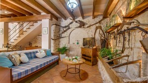 Un interior rústico con vigas de madera a la vista, una pared de piedra, un sofá con ropa de cama escocesa, una mesa de madera y una escalera.