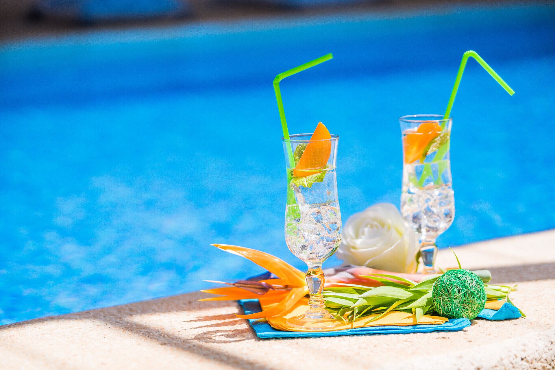 Two tall glasses with straws, filled with a clear liquid and garnished with orange slices and mint leaves, placed on a blue mat beside a pool.