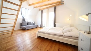 Une chambre avec un plancher en bois, un lit avec une literie blanche, une table de chevet avec une lampe, une fenêtre avec des rideaux et une échelle en bois.