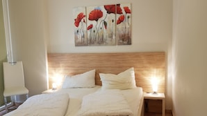 Ein Doppelbett mit zwei weißen Kissen und einem Holzkopfende. Ein Nachttisch mit Lampe und einem Gemälde von roten Blumen hängt über dem Bett.