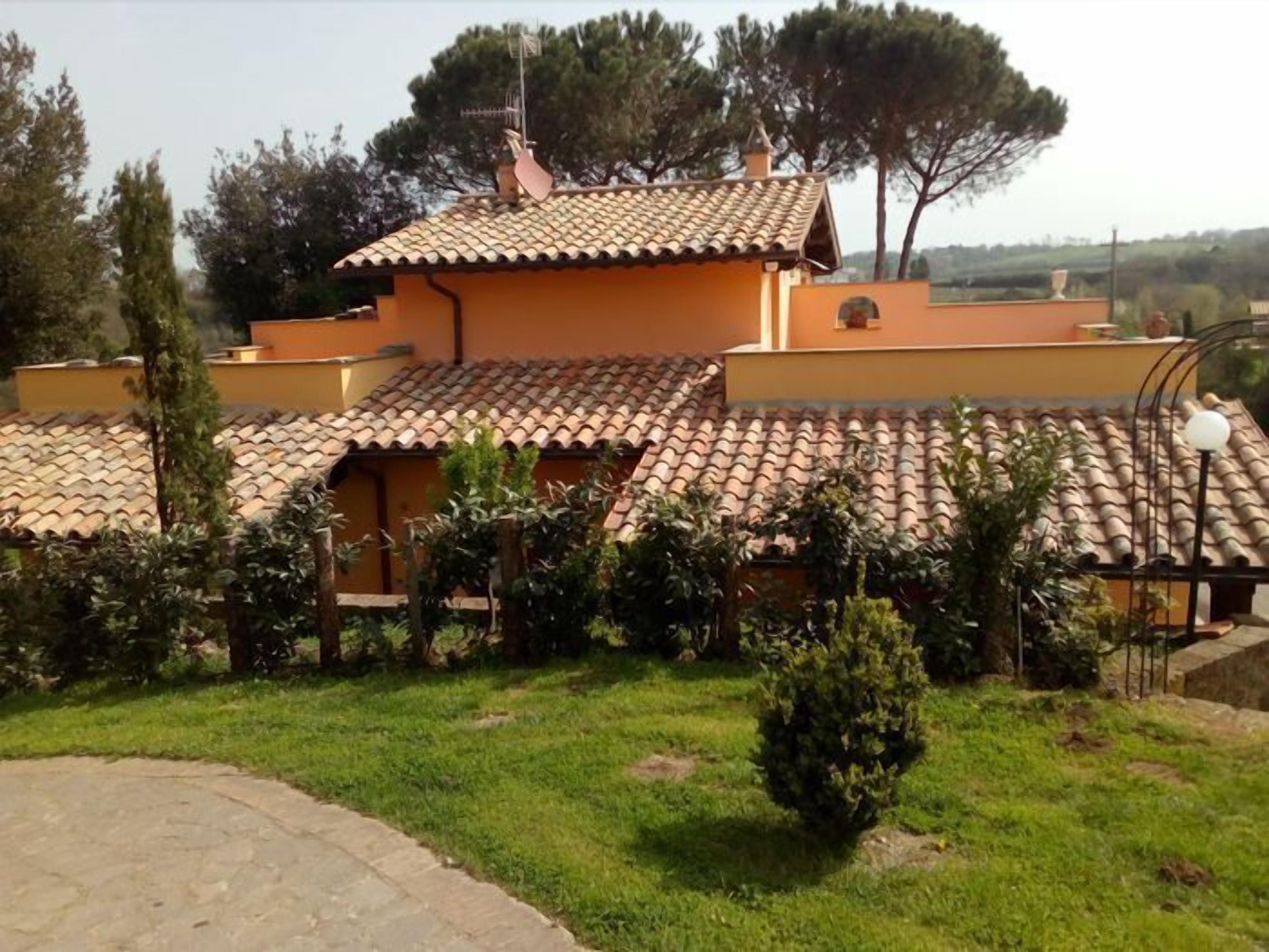 A villa with a tiled roof, surrounded by greenery and a paved area.