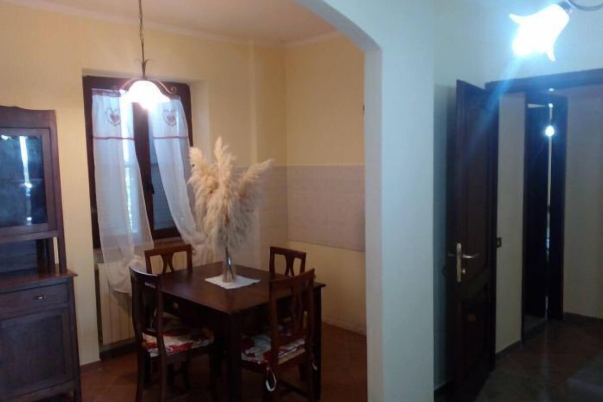 Un comedor con mesa de madera, sillas, un florero con plantas secas y una lámpara colgante.