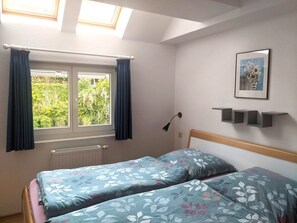 A bedroom with two single beds, a skylight, a window with curtains, a radiator, a wall-mounted lamp, and a framed picture.