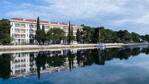 A waterfront hotel with a reflection in the calm water, featuring a multi-story building with balconies and a red roof. - Brijuni Rooms Karmen (Pula)
