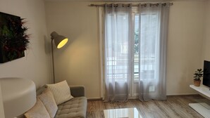 En moderne stue med sofa, gulvlampe, fjernsyn på et stativ og lette gardiner.