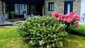 A stone building with a tiled roof, a covered patio with chairs, and a garden with pink flowers. - Casa Garea (Arzua)