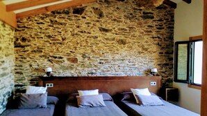 A room with three beds aligned against a stone wall, wooden beams on the ceiling, and a window on the right. - Casa Garea (Arzua)