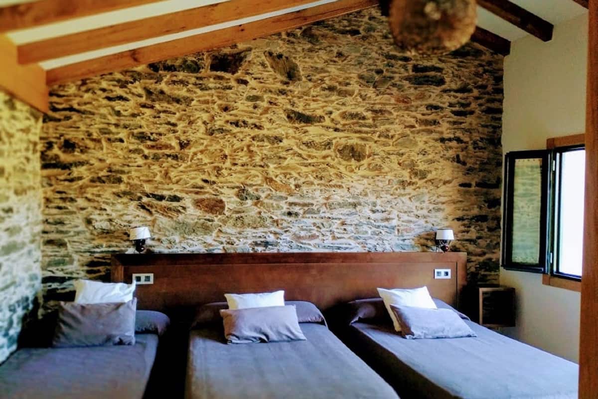 A room with three beds aligned against a stone wall, wooden beams on the ceiling, and a window on the right.