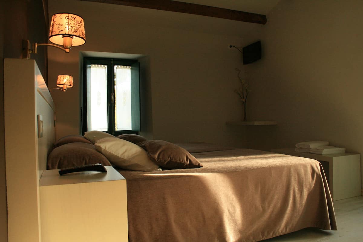 A bedroom with a bed, bedside table, lamp, window, and a small shelf.