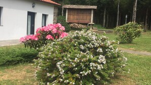 A white building with a red-tiled roof, a wooden structure, and a garden with pink and white flowers. - Casa Garea (Arzua)