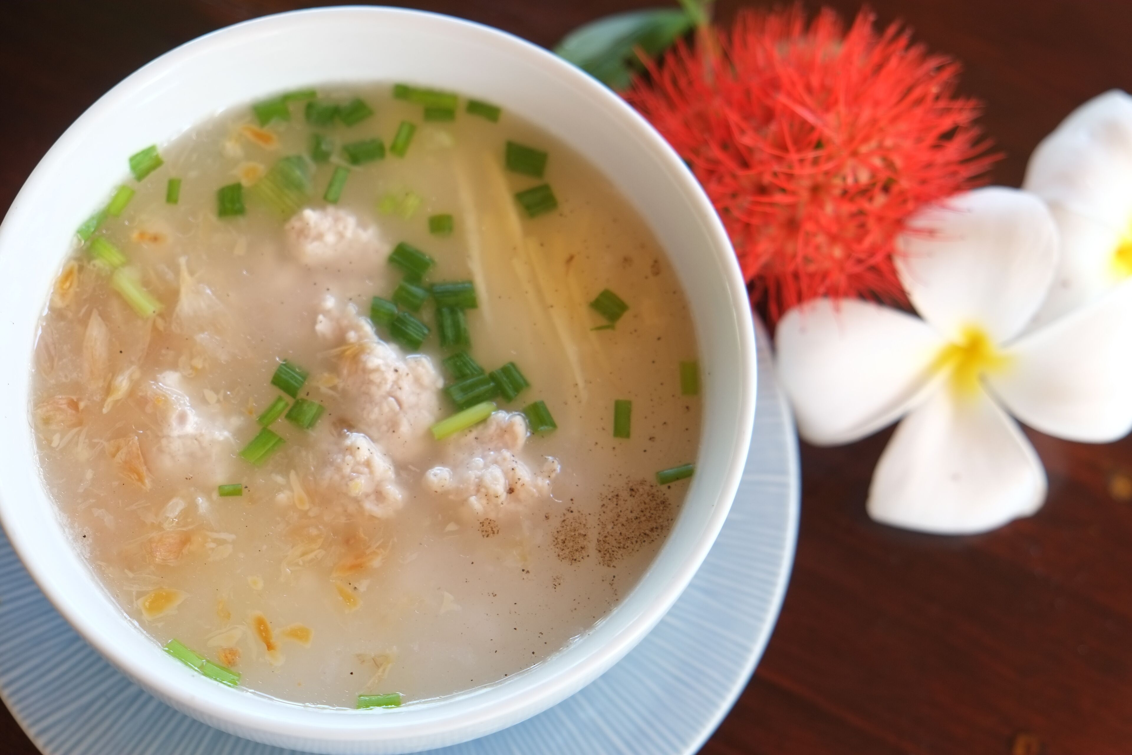 A bowl of soup with green onions and a red flower beside it.