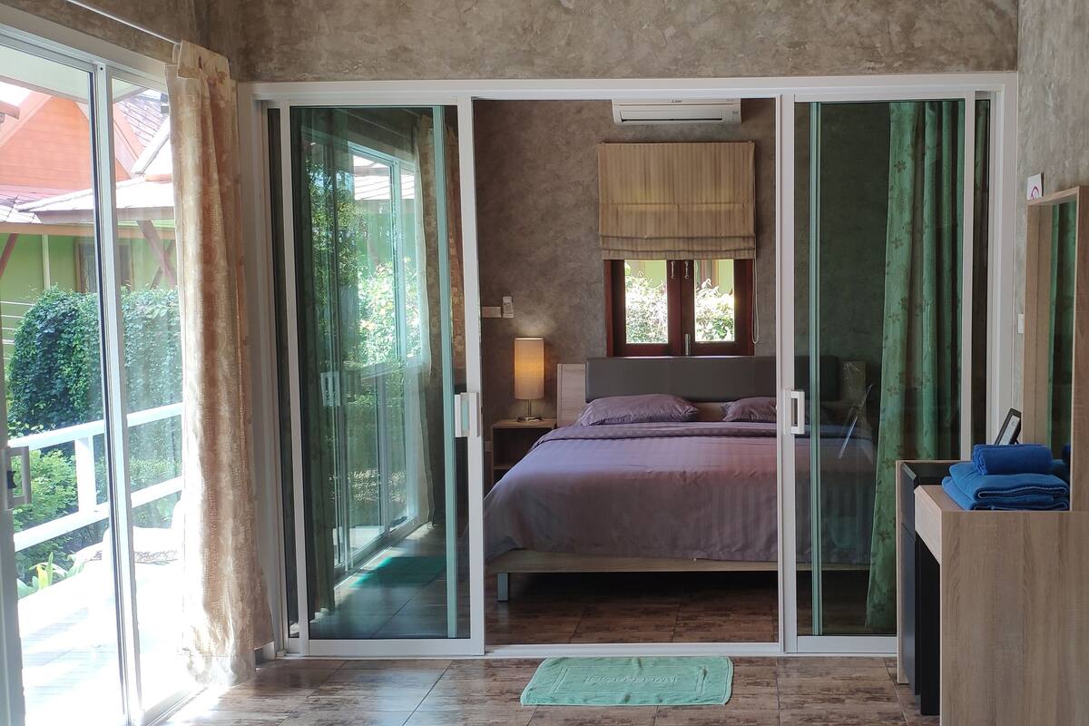 Un dormitorio con una puerta corrediza de vidrio que da a un balcón, una cama con colcha púrpura, una mesita de noche con lámpara y un escritorio de madera con una toalla azul.