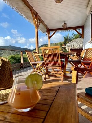 Eine Holzterrasse mit Sitzgelegenheiten im Freien, ein Gebäude mit einem Reetdach und Blick auf Hügel.
