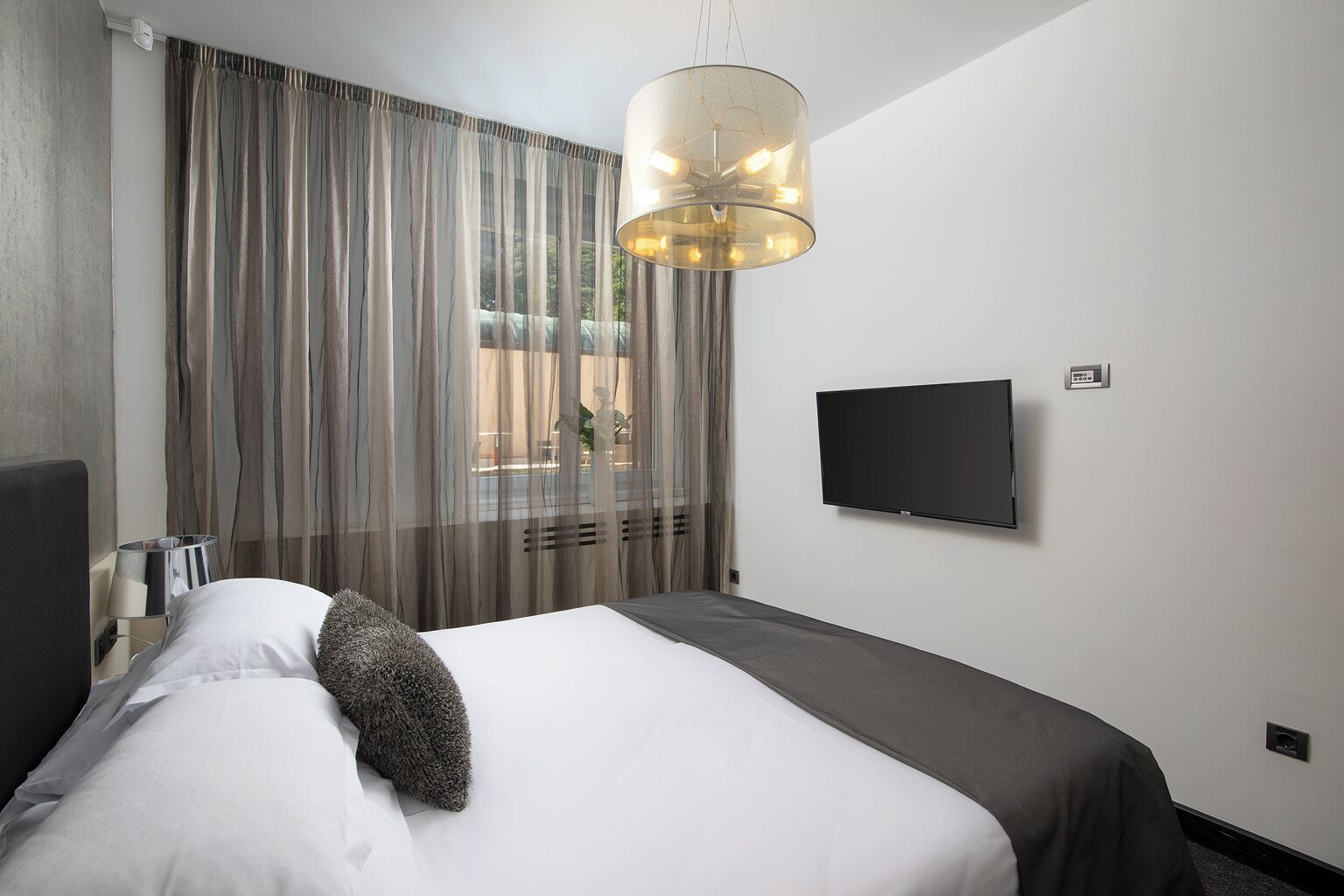 Una camera d'albergo con un letto, una televisione, una finestra con tende e un lampadario.