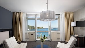 A hotel room with a large window, two white upholstered chairs, a round black table, a vase with flowers, a ceiling light, and a TV on the wall. - Brijuni Hotel Istra (Pula)