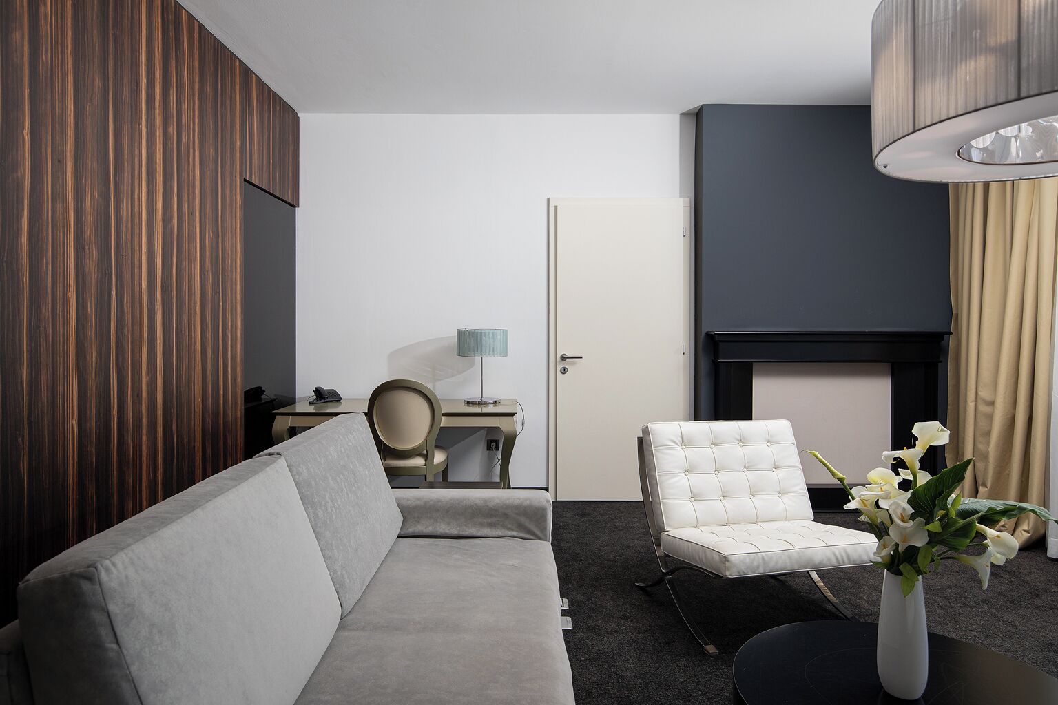 Una habitación de hotel moderna con un sofá gris, un sillón blanco, un escritorio pequeño con una lámpara y un florero con flores.