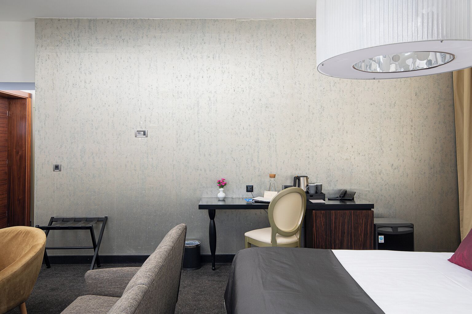A hotel room with a bed, a desk with a chair, a small table with a vase, a lamp, and a wooden door.