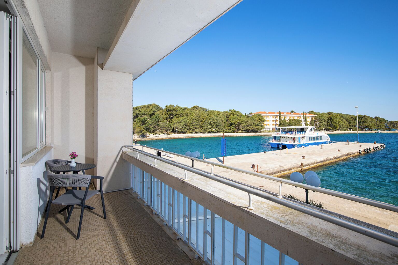 A balcony with a table and chairs, a view of the sea, a dock with a boat, and a building in the background.