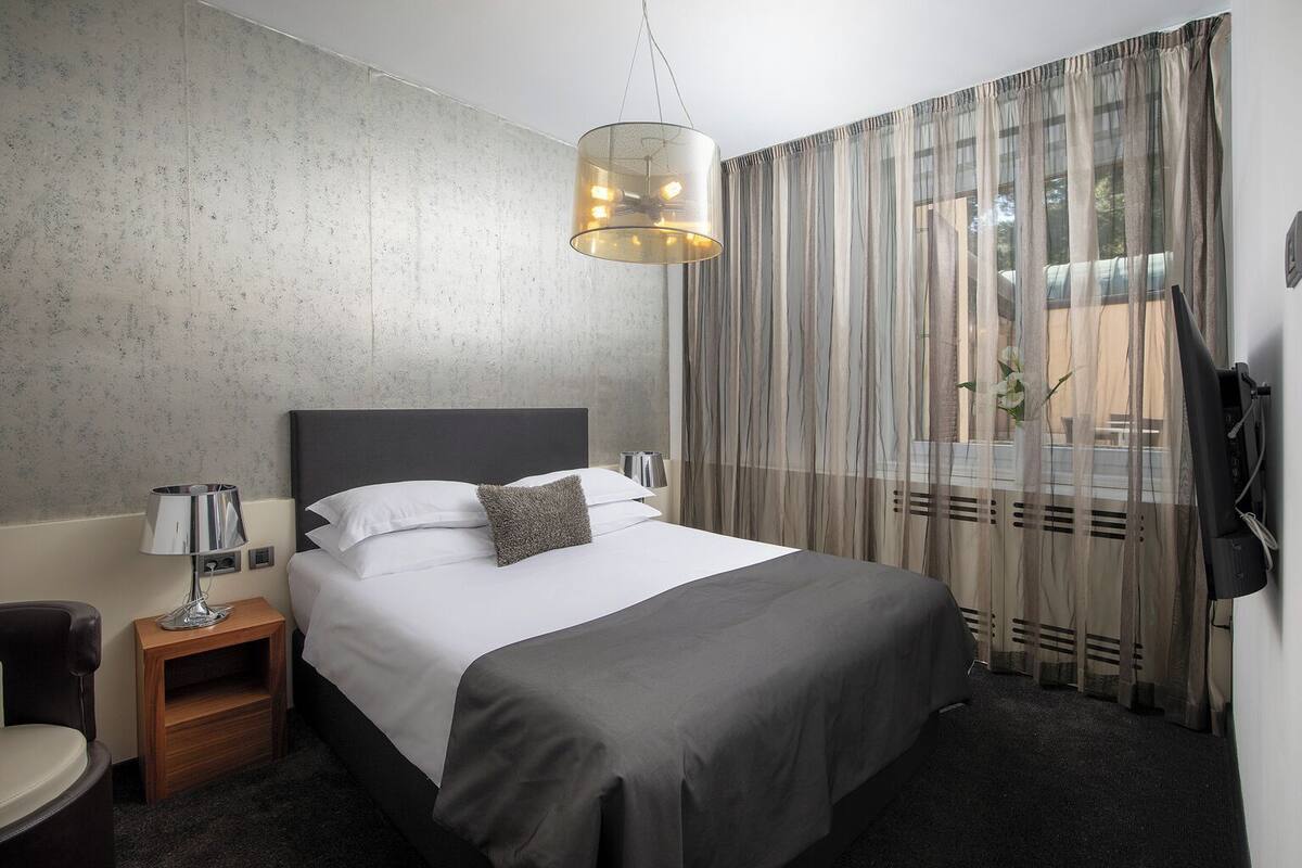 A hotel room with a bed, a nightstand, a lamp, a chair, and a view of the outdoors through sheer curtains.