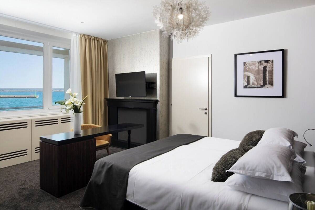 A hotel room with a bed, a desk, a television, a chandelier, a framed picture, and a window with a view of the sea.