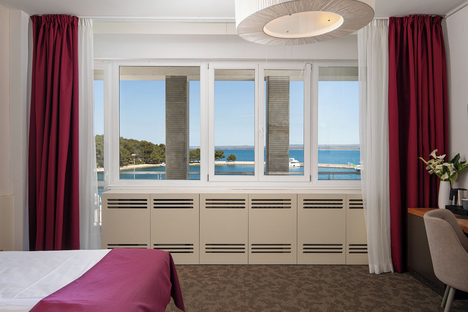 A hotel room with a large window, a bed, a desk, and a chair.