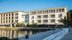 A waterfront hotel with multiple balconies, a pool, and a row of white benches along the water's edge. - Brijuni Hotel Istra (Pula)