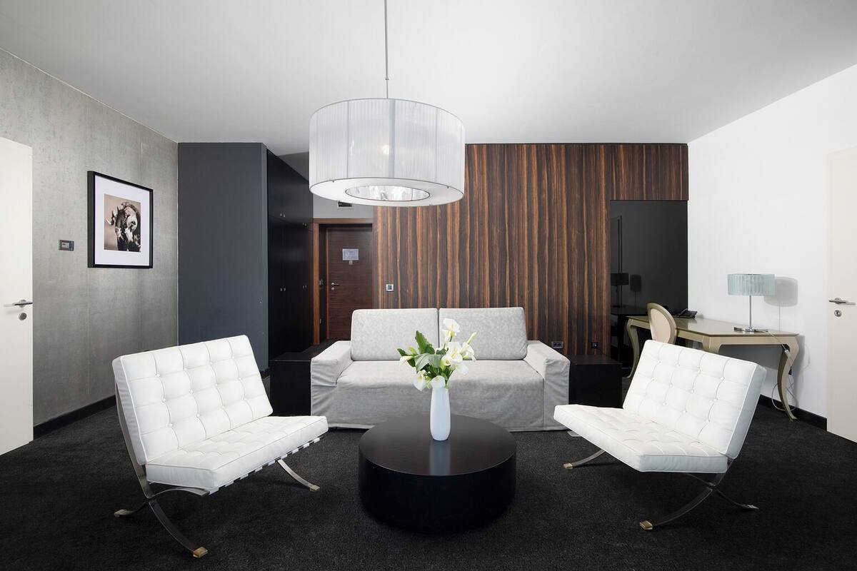 A modern hotel room with a seating area featuring two white upholstered chairs, a round black coffee table, and a clear pendant light.