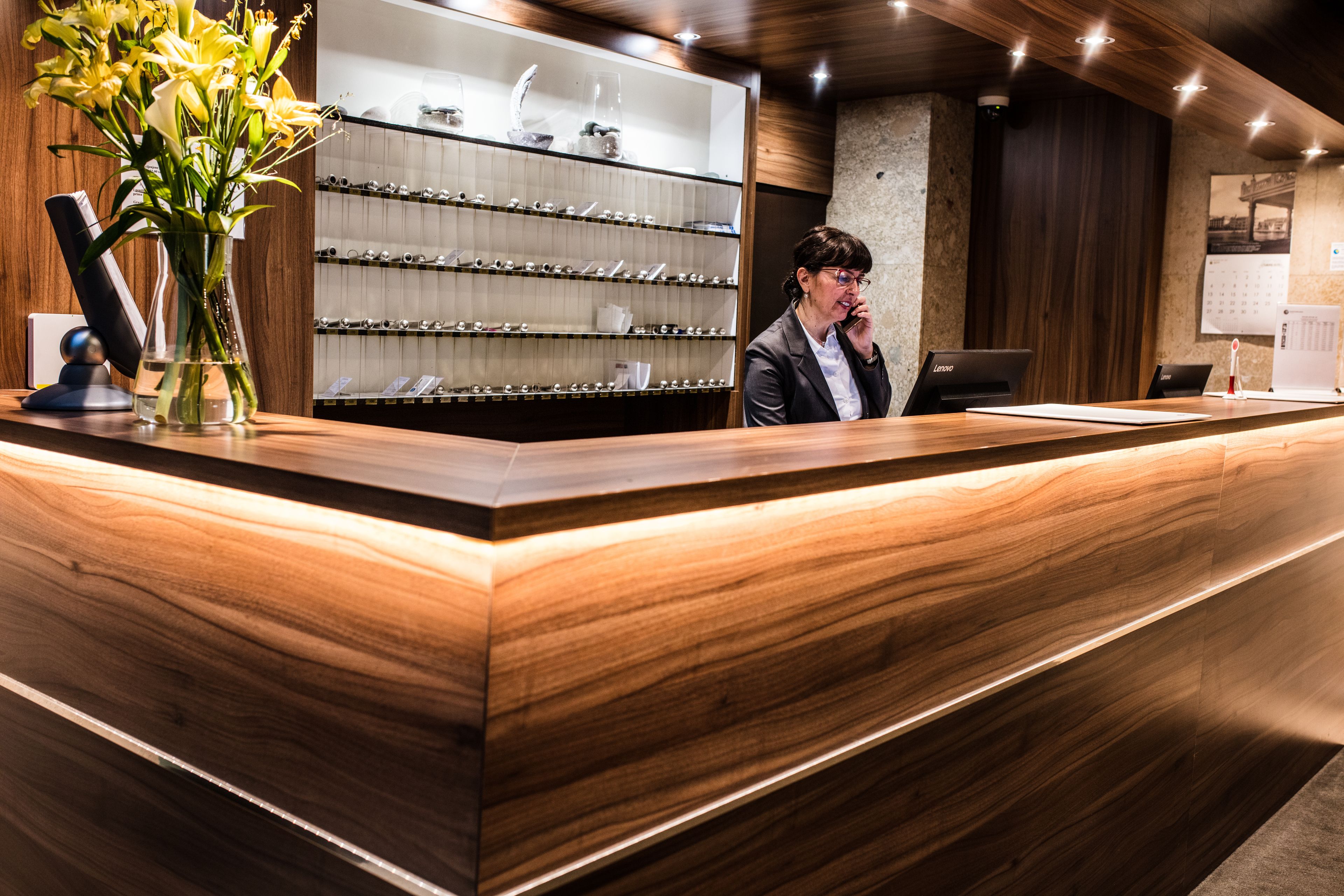 A reception desk with a wooden finish, a vase of yellow flowers, and a display of various glassware.