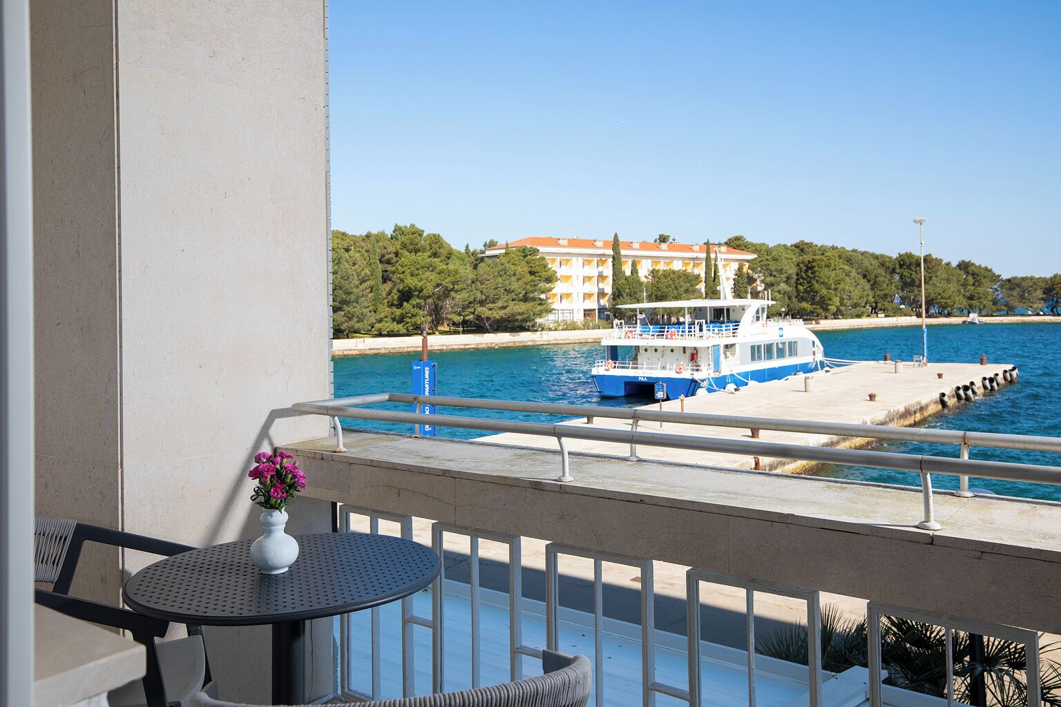 A balcony with a table and chairs, overlooking a body of water with a boat docked at a pier, and a building with a red roof in the background.