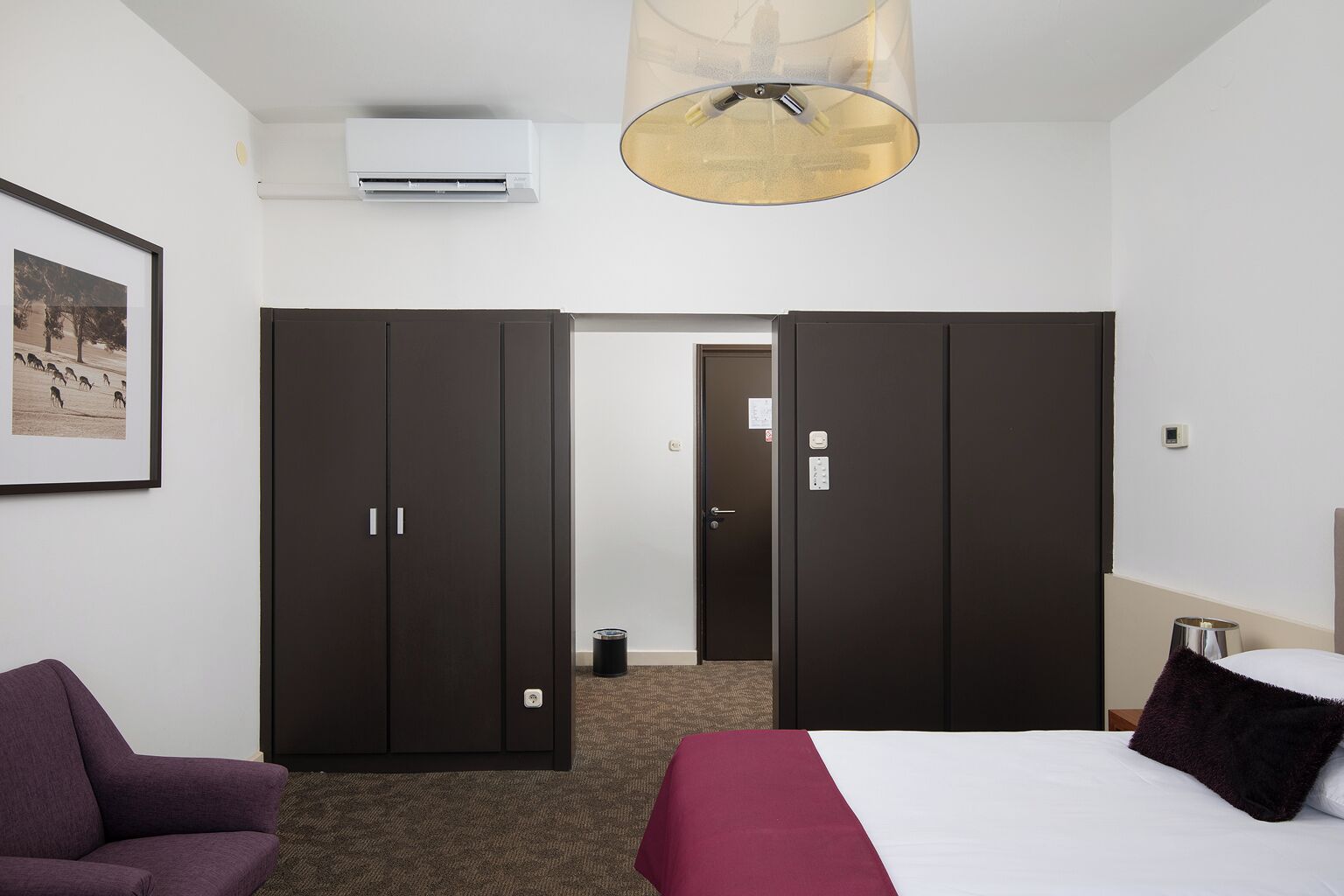 Quarto de hotel com cama, poltrona, guarda-roupa e ar condicionado.