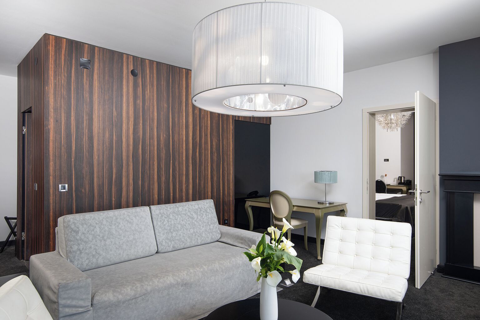 Een moderne hotelkamer met een grijze bank, een witte fauteuil, een eettafel en een grote kroonluchter.