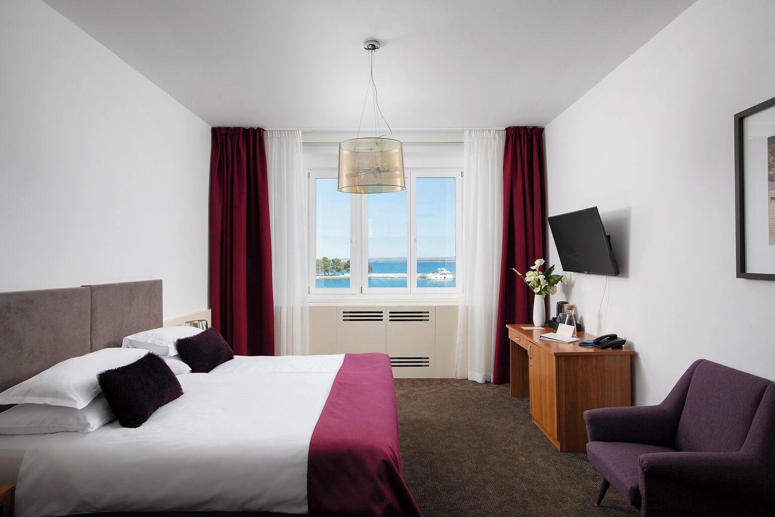 Een hotelkamer met een bed, een bureau, een stoel, een televisie en een raam met uitzicht op zee.