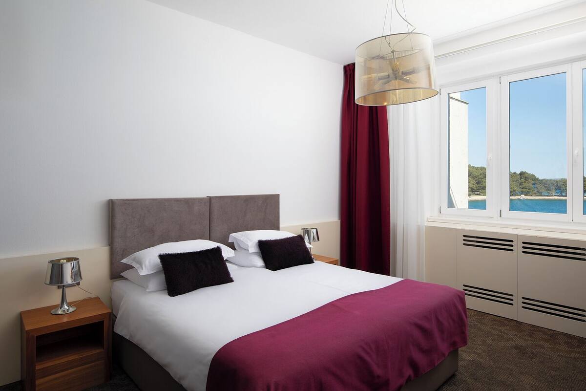 A hotel room with a bed, a nightstand, a lamp, a window with a view, and a ceiling light.
