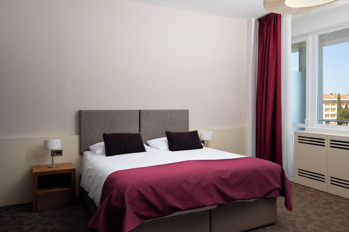 A hotel room with a bed, a nightstand, a window with curtains, and a radiator.
