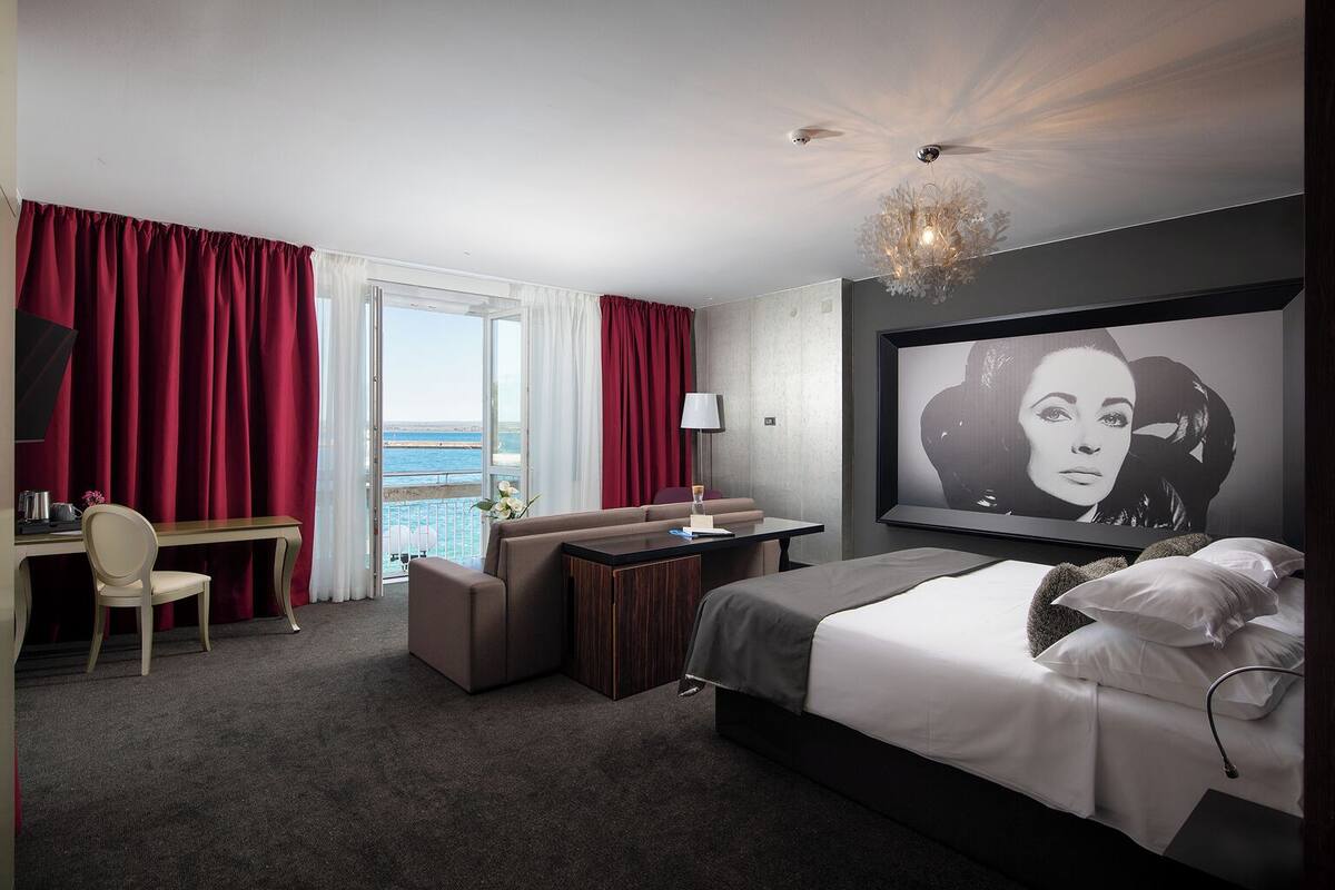A hotel room with a bed, a desk, a chair, a sofa, a TV, and a view of the sea.