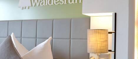 A hotel room with a padded headboard, a bed with white and gray pillows, and a wall-mounted lamp.