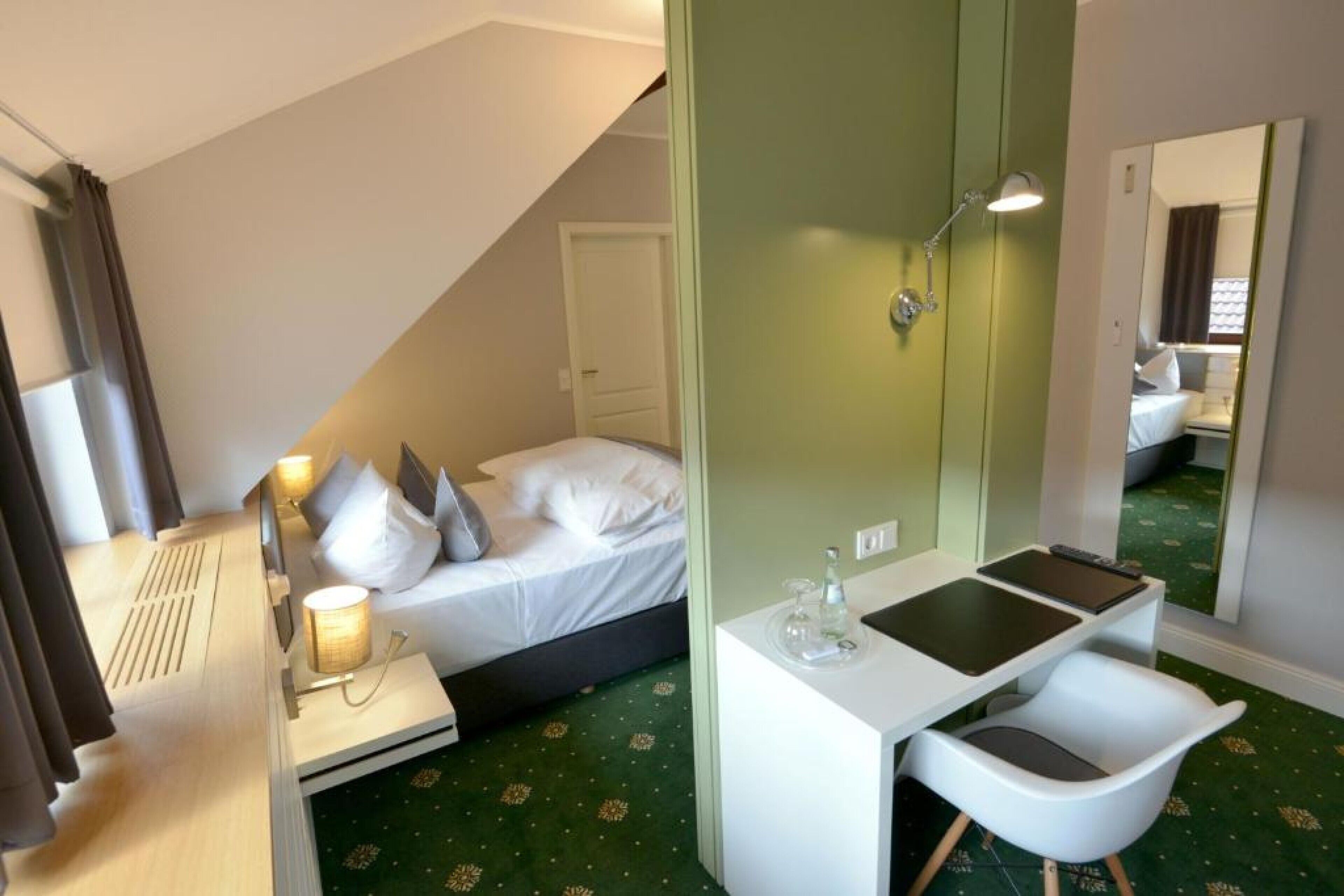 A hotel room with a bed, a desk with a chair, a mirror, and a small table with a lamp.