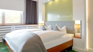 A hotel room with a large bed, a headboard with a logo, a window with curtains, and a radiator below the window. - Hotel Restaurant Waldesruh (Emstek)